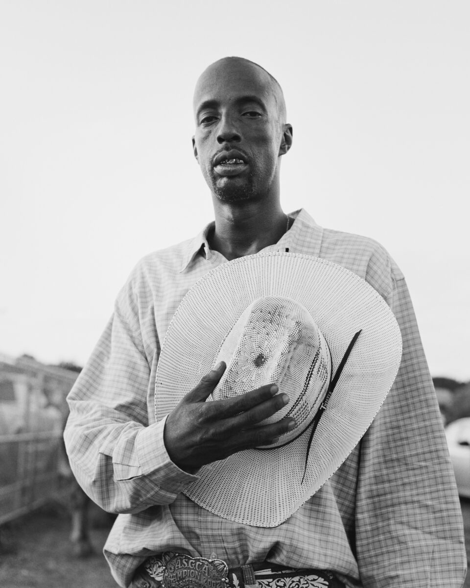 Black and white photograph of a man holding a cowboy hat against his chest while looking at the camera.