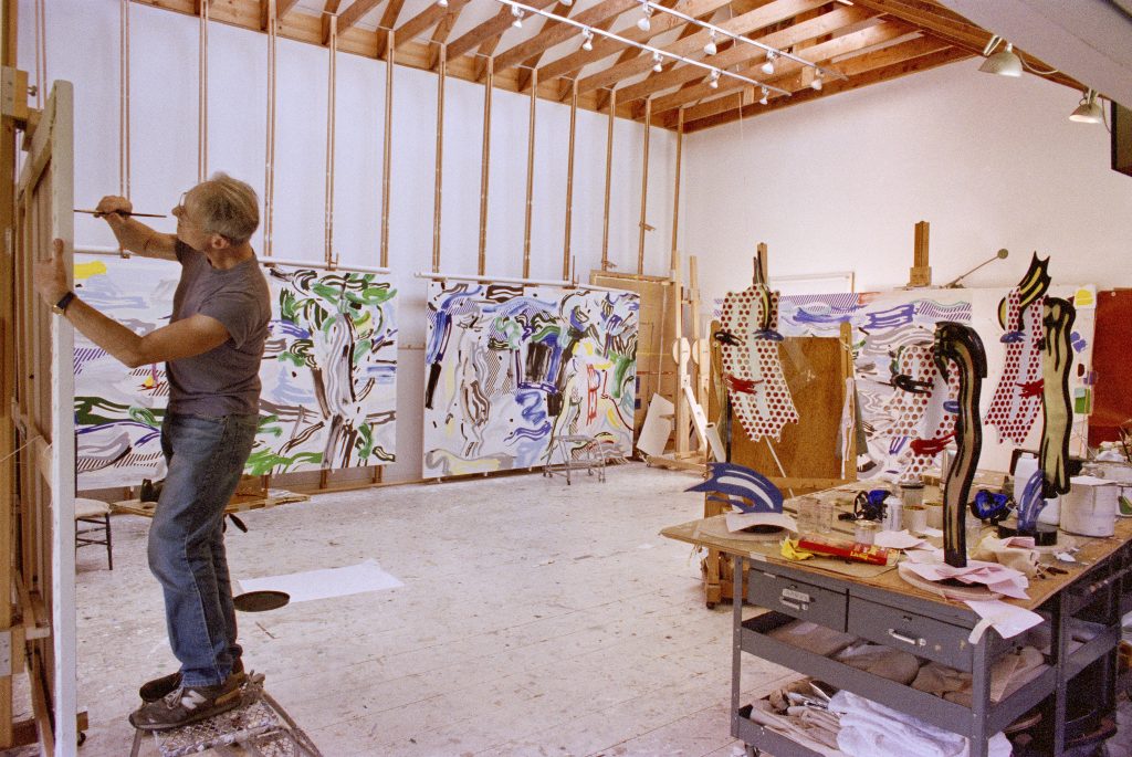 Archival image of Roy Lichtenstein working in his Southampton studio with works featured in the exhibition,1987. Photographed by Bob Adelman. Paintings from left to right: River Scene, 1987; Artemis and Acteon, 1987; Left sculpture: Brushstroke Head I, 1987