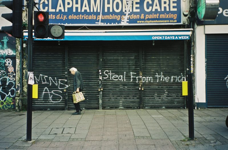 Colour photograph of an elderly person walking along the street past a shuttered shop front that has “STEAL FROM THE RICH” spray-painted in front.