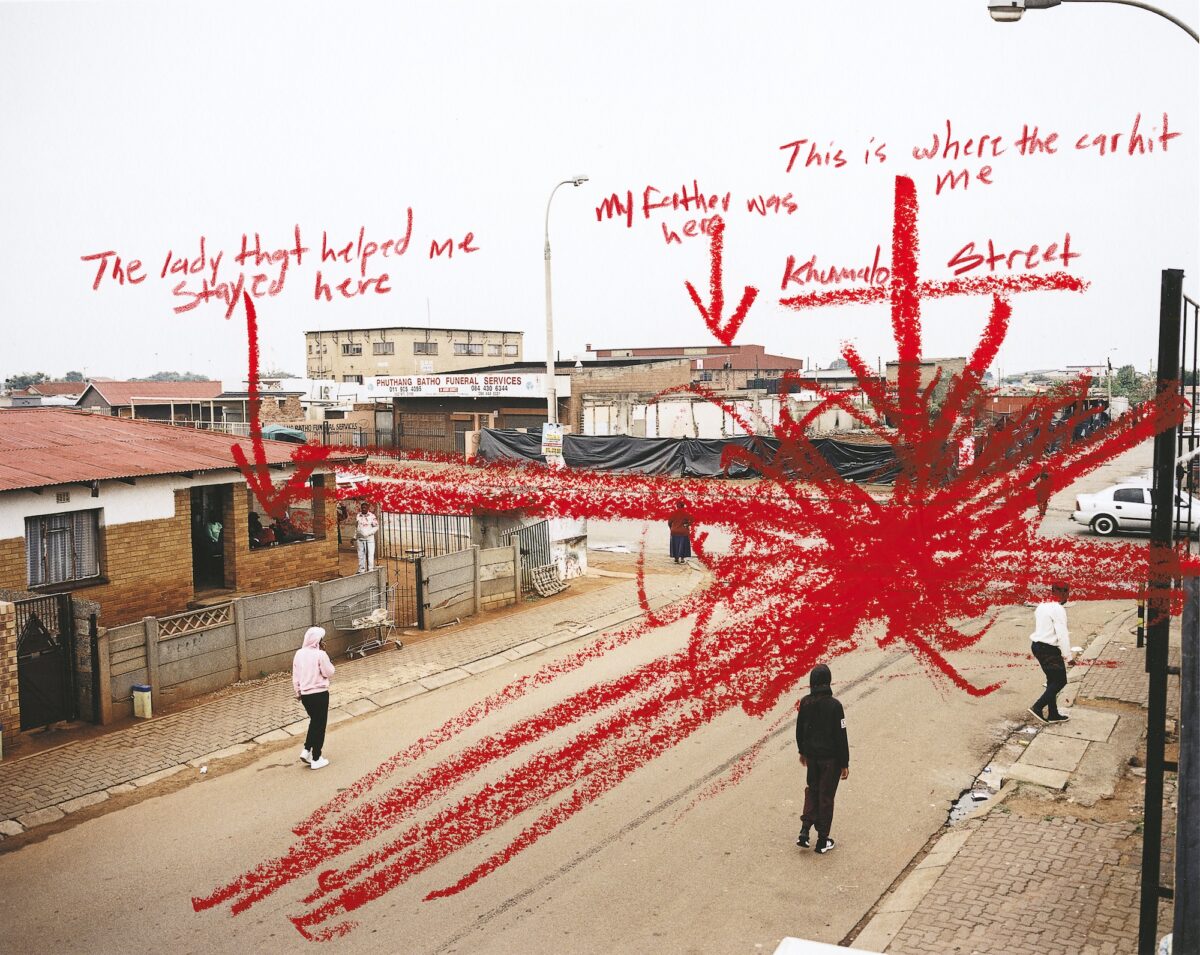 Colour photograph of people stood on a street. The photo is marked with red annotations saying: “The lady that helped me stay here”, “My father was here”, “This is where the car hit me” and “Khumalo Street.”