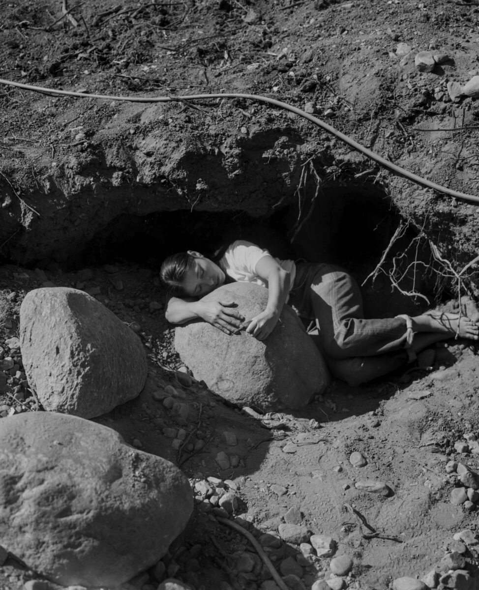 Black and white photograph of a woman curled up around a rock on the ground.